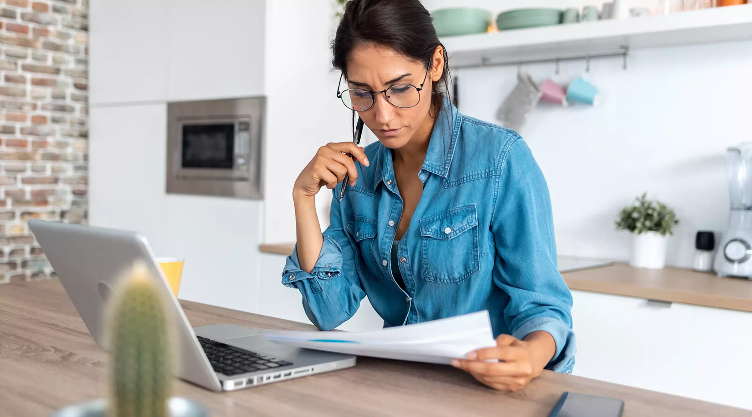 Junge Frau, die Papierkram überprüft und zu Hause an einem Laptop in der Küche arbeitet. Junge Frau, die Papierkram überprüft und zu Hause an einem Laptop in der Küche arbeitet.